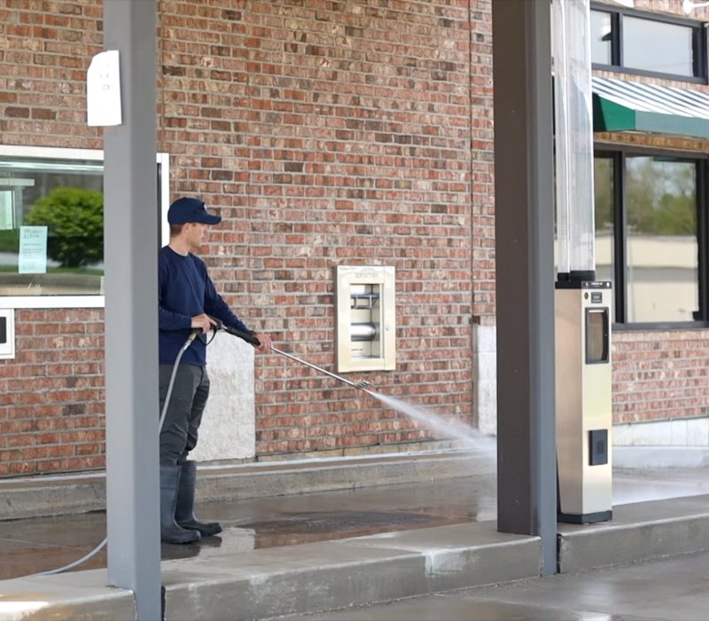 A worker uses a pressure washer for commercial concrete cleaning outside a brick building.